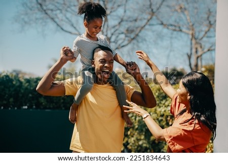 Father carrying his daughter on his shoulders while standing outside with mom. Happy parents playing with their kid at home. Family laughing and having fun in the sun.