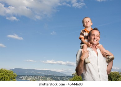 Father Carrying His Son On Piggy Back Ride Outdoors Against Nature And Blue Sky