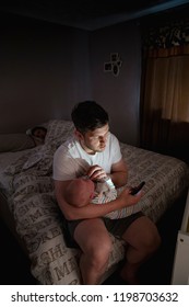 Father Checking On His Newborn Baby Boy At Night As His Wife Stays Asleep In Bed. While Waiting On The Baby To Fall Asleep He Is Looking At His Mobile Phone.