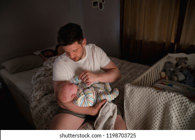 Father Checking On His Newborn Baby Boy At Night As His Wife Stays Asleep In Bed.