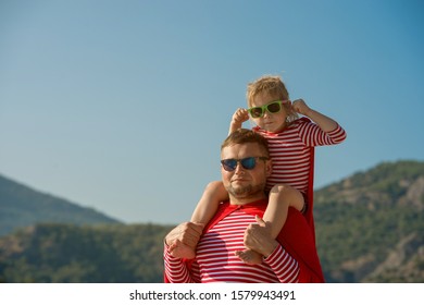 Father And Child Girl Playing Superhero. Superman Dad And Daughter Having Fun. Imagination And Dreams Of Being Hero. Child In Red Cloak, Striped Shirt And Sunglasses On Dads Back. 