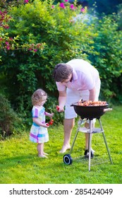 Father And Child Grilling Meat. Family Camping And Enjoying BBQ. Dad And Daughter At Barbecue Preparing Steaks And Sausages. Parents And Kids Eating Grill Meal Outdoors. Garden Fun For Children. 