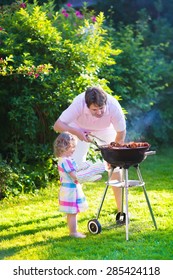 Father And Child Grilling Meat. Family Camping And Enjoying BBQ. Dad And Daughter At Barbecue Preparing Steaks And Sausages. Parents And Kids Eating Grill Meal Outdoors. Garden Fun For Children. 