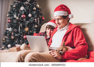 Father With Child And Puppy Dog In Santa Hats Having A Video Call On Christmas Day At Laptop, Sitting On A Couch In The Living Room With Christmas Tree At Home