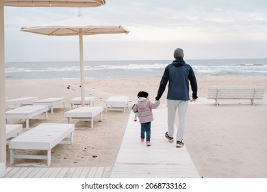 Father with child walking to the beach in autumn fall holding hands, offseason sea. Fresh air, healthy lifestyle, family spending time outdoors. Toddler girl with sporty man on empty shore