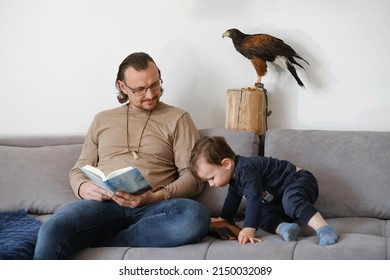 Father With Child And Wild Bird Buzzard Sitting On The Coach, Reading, Playing On Smartphone. Fatherhood, Unusual Family With Pet At Home. Extraordinary Animal Friend