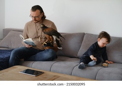 Father With Child And Wild Bird Buzzard Sitting On The Coach, Reading, Playing On Smartphone. Fatherhood, Unusual Family With Pet At Home. Extraordinary Animal Friend