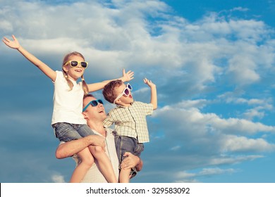 Father And Children Playing In The Park  At The Day Time. Concept Of Friendly Family. Picture Made On The Background Of Blue Sky.