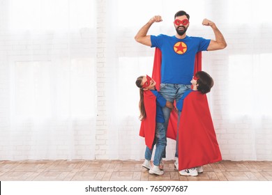 Father with children in red and blue suits of superheroes. On their faces are masks and they are in raincoats. They are posing in a bright room.