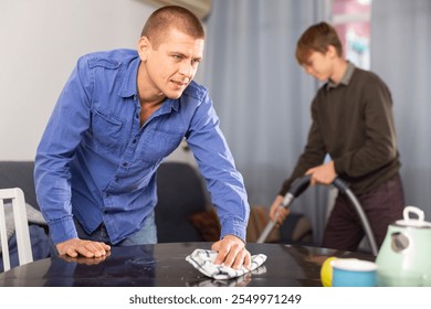 Father cleans the table with a rag while son vacuums the room - Powered by Shutterstock