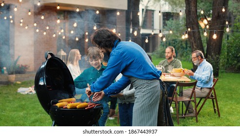 Father Cooking Food On Barbecue For Weekend Lunch And Giving Hot Meat And Corn To Small Cheerful Kids. Family At Picnic On Back Yard. Little Son And Daughter At Dad Waiting As Man Preparing Eating.