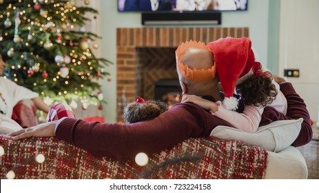 Father Is Curled Up On The Sofa With His Daughter At Christmas Time, Watching Television.