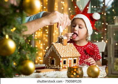 Father and cute child in red hat building gingerbread house together. Girl licking glaze off. Beautiful decorated room with lights and Christmas tree, candles and lanterns. Happy family celebrating.