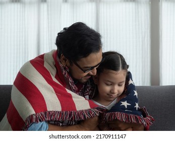 Father and cute girl sitting close to each other wrapped in big rug with design of United States of America flag. Daddy and daughter celebrating 4th of July, national holidays.