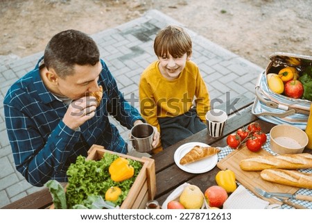 Father dad and school kid boy child eating croissant picnic forest camping site with vegetables, juice, coffee, and croissants. Wooden crate with fresh organic veggies surrounded with bread baguettes