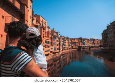 A father and daughter admire the calm river in Girona, Spain, during the flower festival, with a blue sky and reflections of colorful buildings in the water - Powered by Shutterstock