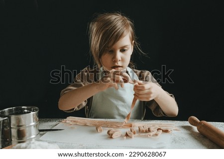 Father and daughter in aprons holding dough cut into strips on a white table against a dark wall. Parent with child preparing homemade pastries or noodles. Selective focus.
