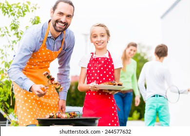 Father And Daughter Barbecue Meat Spits And Sausages At Family Garden BBQ 