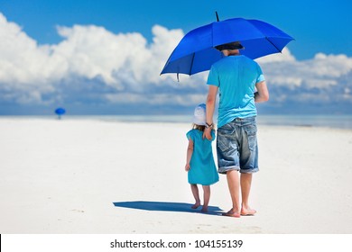 Father And Daughter At Beach With Blue Umbrella To Hide From The Sun