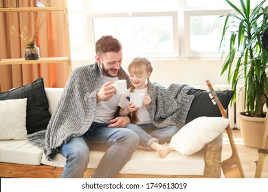 Father And Daughter. Handsome Young Man And Little Cute Girl Sitting On The Sofa At Home, Covered Themselves With A Warm Blanket, Holding White Cups With Hot Tea On A Sunny Winter Day. Father's Day.