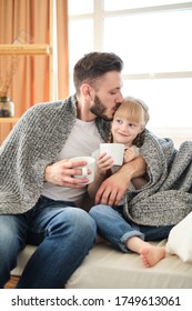 Father And Daughter. Handsome Young Man And Little Cute Girl Sitting On The Sofa At Home, Covered Themselves With A Warm Blanket, Holding White Cups With Hot Tea On A Sunny Winter Day. Father's Day.