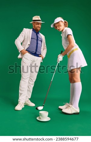 Father and daughter have fun on golf court. Dad in smart casual outfit with cup of coffee and dressed like golf player girl teenager on green background. Concept of parenthood, hobby, recreation.