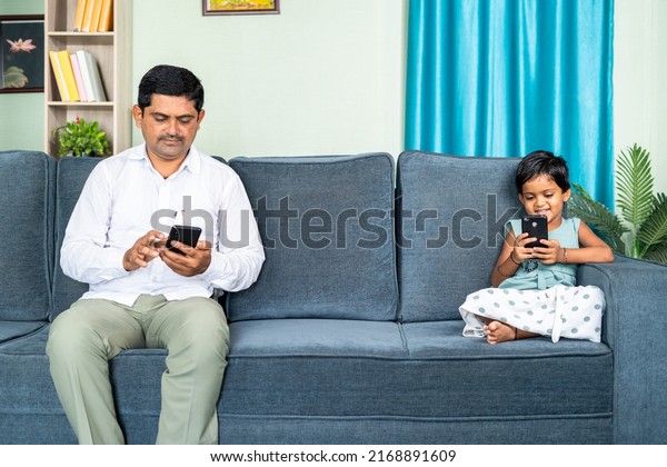 Father and daughter on sofa separately busy\
using mobile phone at home - concept of digital divide, social\
media and technology\
addiction