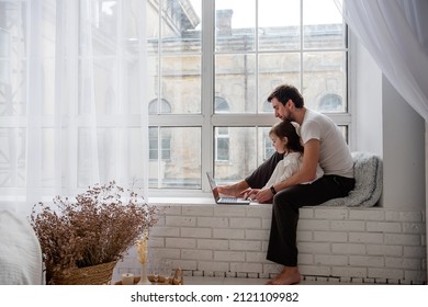 Father And Daughter In Pajamas Are Playing On Laptop, Sitting On Wooden, White Windowsill Near The Window. Young Man Teaches Little Girl How To Work On Computer. Technological Generation Of The Family