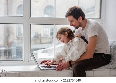 Father and daughter in pajamas are playing on laptop, sitting on wooden, white windowsill near the window. Young man teaches little girl how to work on computer. Technological generation of the family