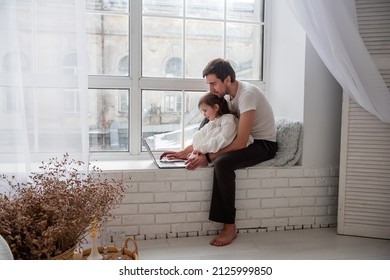 Father and daughter in pajamas are playing on laptop, sitting on wooden, white windowsill near the window. Young man teaches little girl how to work on computer. Technological generation of the family