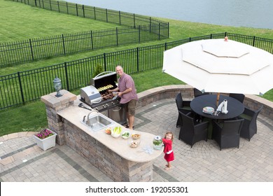 Father and daughter preparing a barbecue at an outdoor summer kitchen on a paved patio with a garden umbrella, table and chairs as they grill the meat on the gas BBQ waiting for guests to arrive