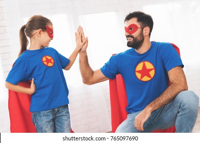 Father with daughter in red and blue suits of superheroes. On their faces are masks and they are in raincoats. They are posing in a bright room.