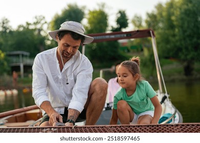 Father and daughter securing a boat to a dock with ropes. They are both kneeling on the boat, and the man is wearing a sun hat and a white shirt. The background shows other boats and a marina. - Powered by Shutterstock