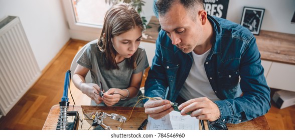 Father And Daughter Sitting By The Wooden Table Holding Circuit Board And Building Robot At Home As A School Science Project