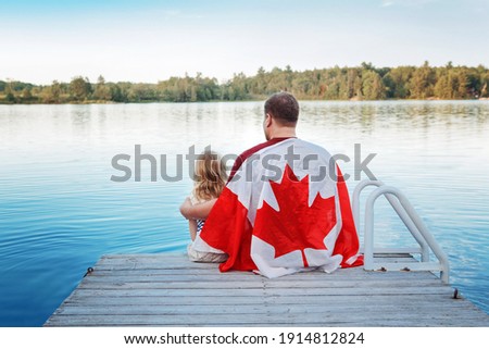 Father and daughter wrapped in large Canadian flag sitting on wooden pier by lake. Canada Day celebration outdoor. Dad and child sitting together on 1 of July celebrating national Canada Day.