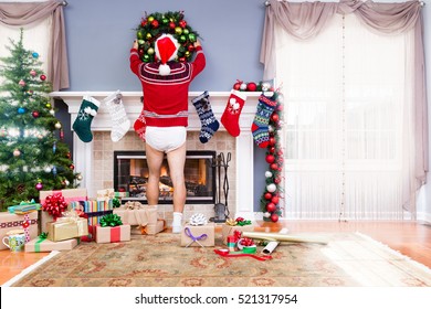 Father Decorating The Living Room For Christmas In His Underpants And Santa Outfit Hanging A Wreath Above The Mantelpiece Alongside Colorful Gifts And A Decorated Tree