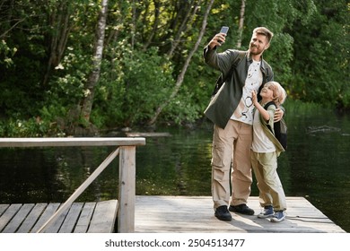 Father engaging in outdoors activity while taking selfie with his son using mobile device on wooden dock by water surrounded by lush greenery - Powered by Shutterstock