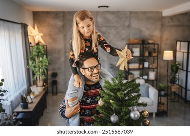 A father with eyeglasses and sweater and daughter a shoulder ride decorate a Christmas tree with ornaments and star inside their cozy, warmly lit home - Powered by Shutterstock