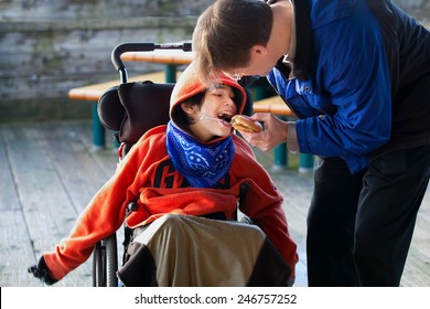 Father Feeding Disabled Son A Hamburger In Wheelchair. Child Has Cerebral Palsy