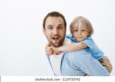 Father Giving Piggyback Ride To Cute Blond Son With Vitiligo. Potrait Of Carefree Beautiful Dad And Kid, Hugging, Showing Tongue And Making Funny Faces While Playing And Fooling Around Over Gray Wall