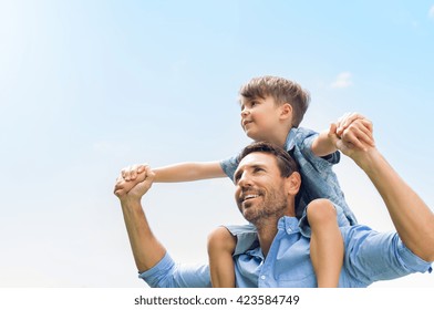 Father giving son ride on back in park. Portrait of happy father giving son piggyback ride on his shoulders and looking up. Cute boy with dad playing outdoor.