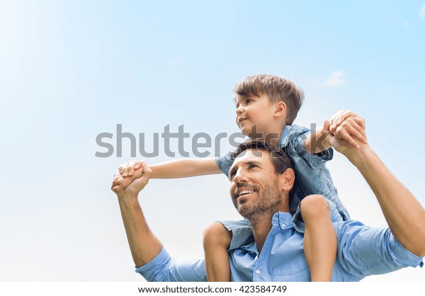 Father giving son ride on back in\
park. Portrait of happy father giving son piggyback ride on his\
shoulders and looking up. Cute boy with dad playing\
outdoor.