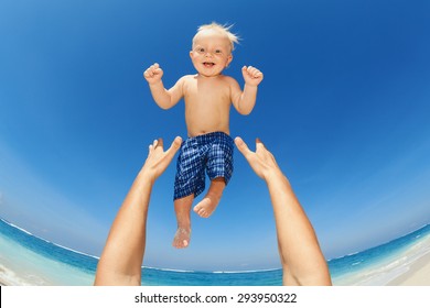 Father Hands Tossing Up The High Air Joyful Baby Boy In On White Sand Sea Beach. Outdoors Healthy Child Activity, Active Lifestyle And Having Fun On Family Summer Vacation With Son On Tropical Island