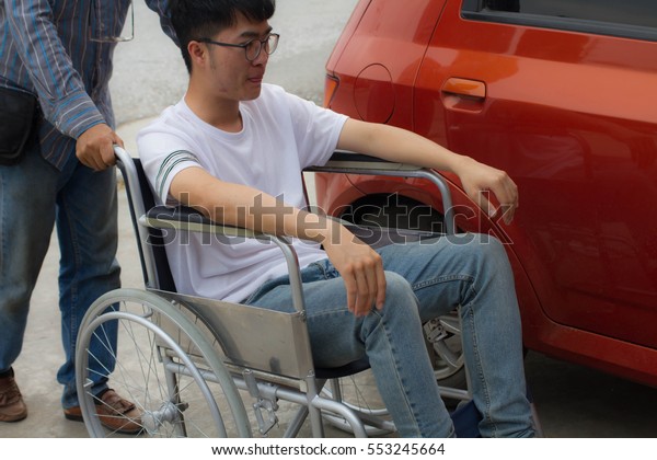 Father helping his\
son who was disabled man wears white t-shirt and blue jeans/ eyes\
glasses  holding the  phone  on wheelchair open the red car near\
the road  in front of\
house