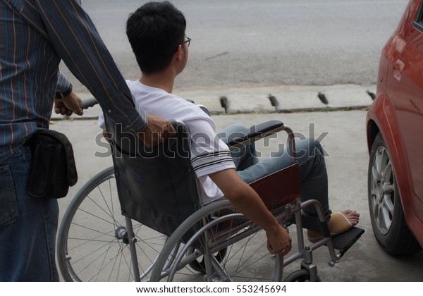 Father helping his\
son who was disabled man wears white t-shirt and blue jeans/ eyes\
glasses  holding the  phone  on wheelchair open the red car near\
the road  in front of\
house