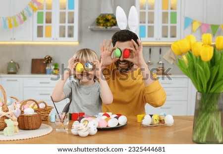 Father and his cute son covering eyes with Easter eggs at table in kitchen