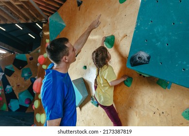 Father And His Daughter Rock Climbers Training In A Modern Indoor Rock Climbing Gym. Male Instructor Helps Little Girl Preschooler On A Private Coach Class. Family Sport Activities. Selective Focus.