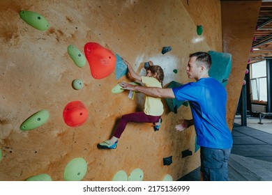 Father And His Daughter Rock Climbers Training In A Modern Indoor Rock Climbing Gym. Male Instructor Helps Little Girl Preschooler On A Private Coach Class. Family Sport Activities. Selective Focus.