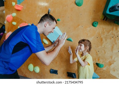 Father and his daughter rock climbers training in a modern indoor rock climbing gym. Male instructor giving high five to little girl preschooler on a private coach class. Family sport activities.  - Powered by Shutterstock