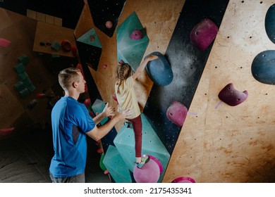 Father And His Daughter Rock Climbers Training In A Modern Indoor Rock Climbing Gym. Male Instructor Helps Little Girl Preschooler On A Private Coach Class. Family Sport Activities. Selective Focus.